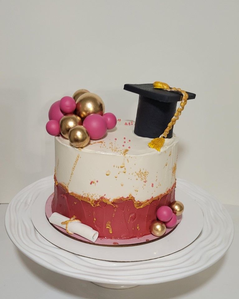 A graduation-themed cake with pink and gold décor, topped with a black graduation cap and a diploma scroll, on a white cake stand.